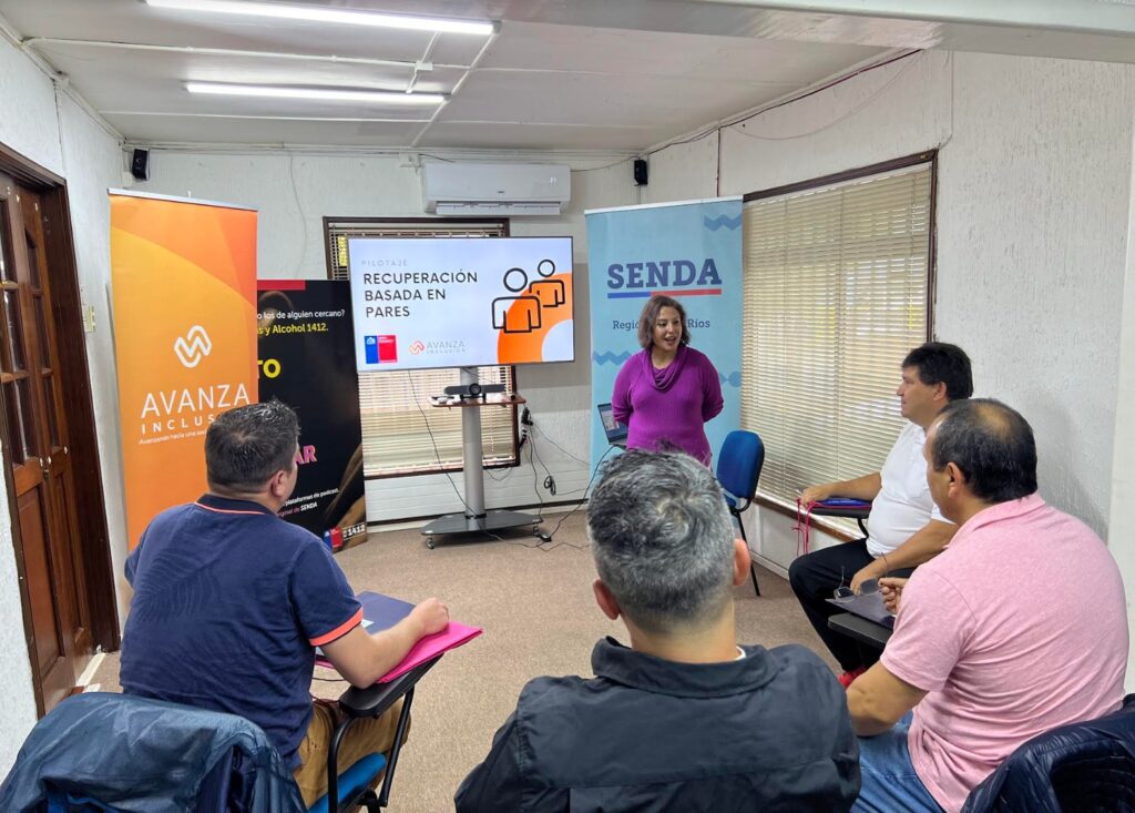 En la sala de presentación, cuatro hombres están sentados de espaldas, enfocados en una mujer que se encuentra de pie frente a ellos. La mujer está dando una presentación sobre el programa "Recuperación basada en Pares". En la pantalla o pizarra de la sala, se visualiza el título del programa. El ambiente sugiere un momento de atención y compromiso hacia la propuesta que se está compartiendo.