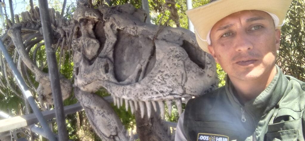 Participante egresado posando a la foto en su vestimenta laboral junto a un esqueleto de dinosaurio, correspondiente a su lugar de trabajo, Buin Zoo.